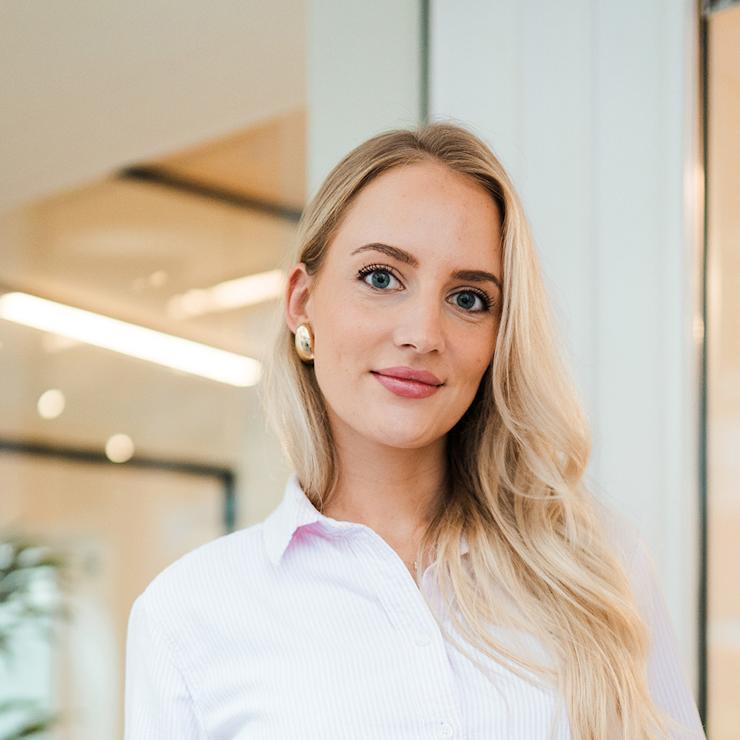 Junge Frau mit langen blonden Haaren und Brille steht in einem modernen Innenraum.