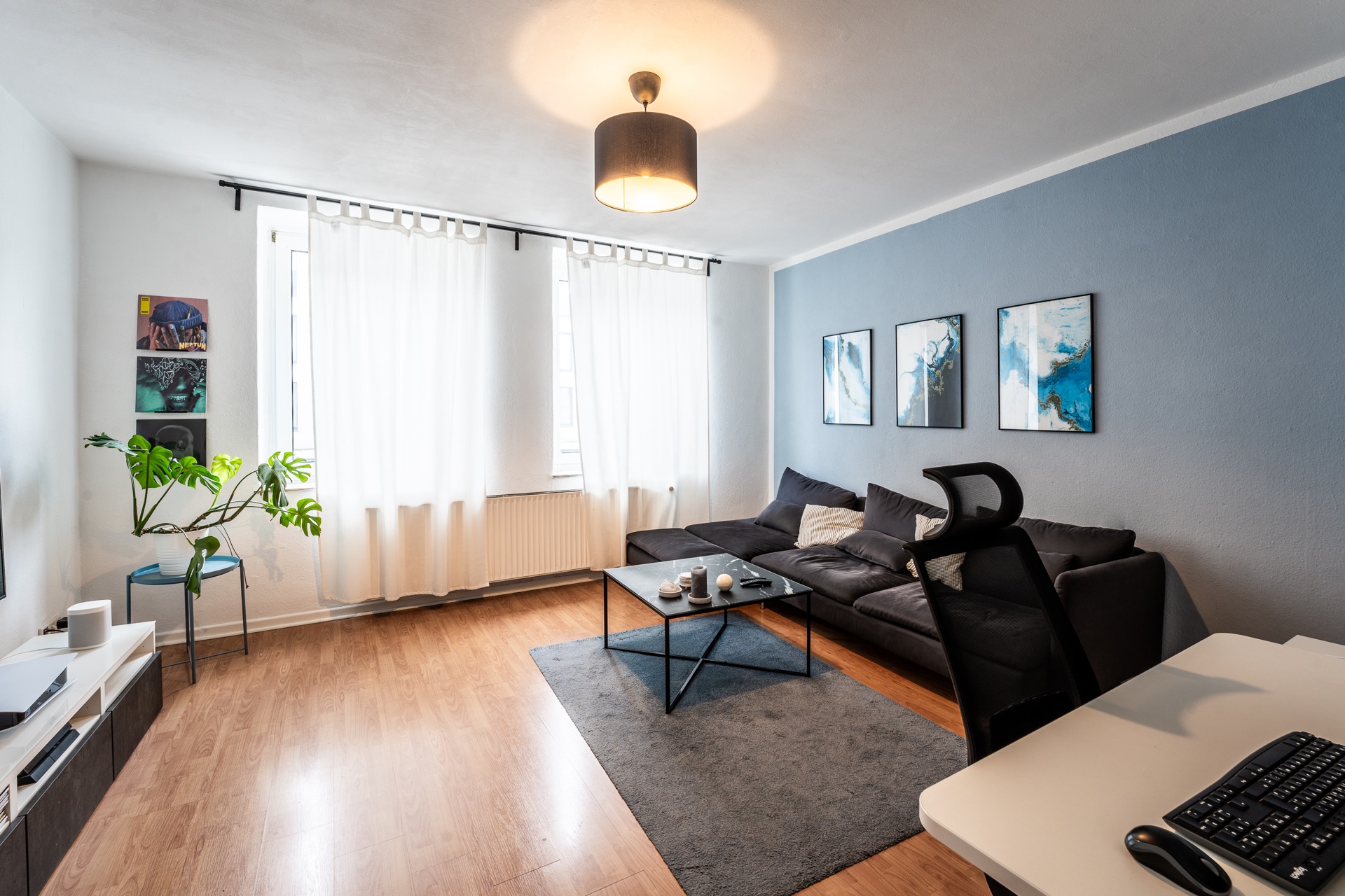 Helles Wohnzimmer mit Sofa, Couchtisch, Teppich, Schreibtisch und großen Fenstern mit weißen Vorhängen.