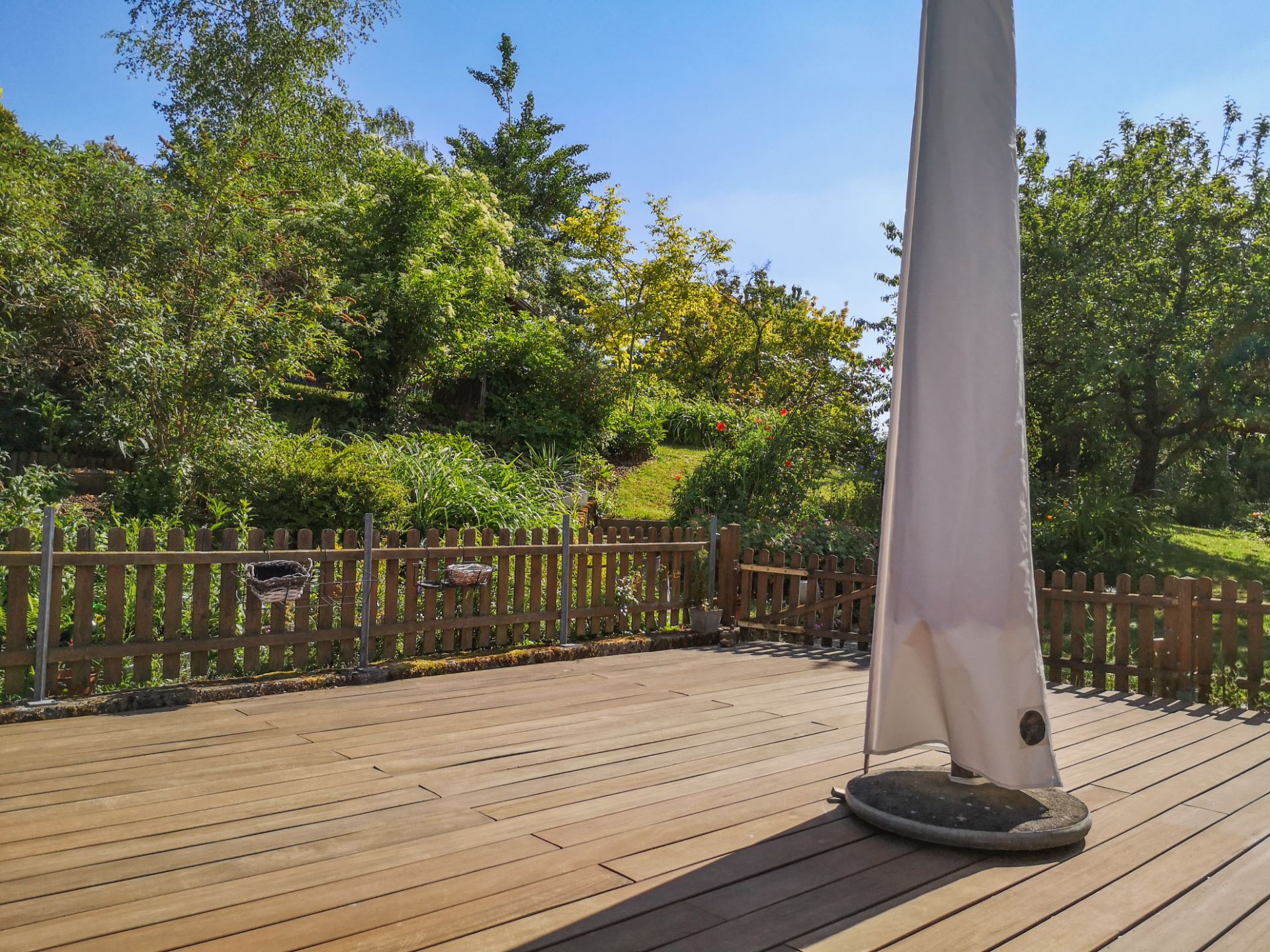 Holzterrasse mit geschlossenem Sonnenschirm, dahinter ein Garten mit Bäumen und Holzzaun.