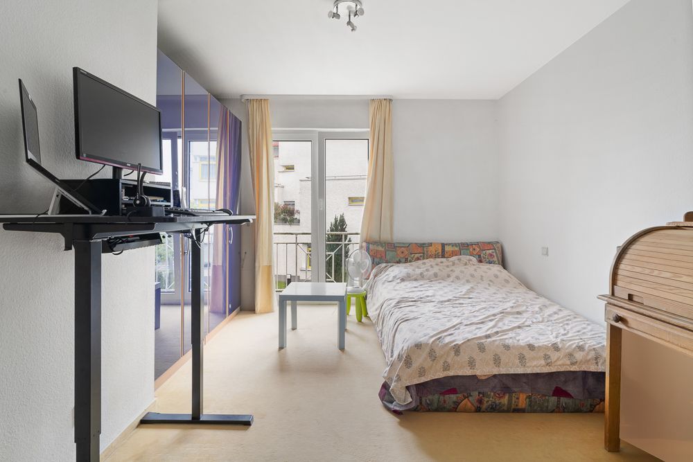 Helles Schlafzimmer mit Bett, Schreibtisch, Stehpult und Zugang zu einem Balkon.