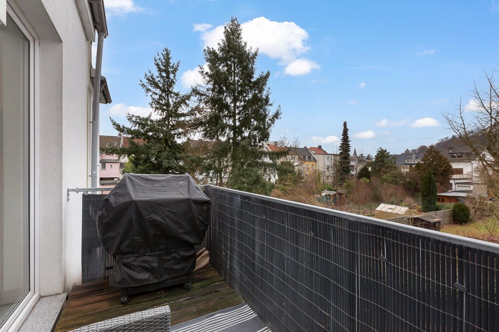 Balkon mit Holzboden, Grill mit Abdeckung und Ausblick auf Nachbarhäuser und Bäume.