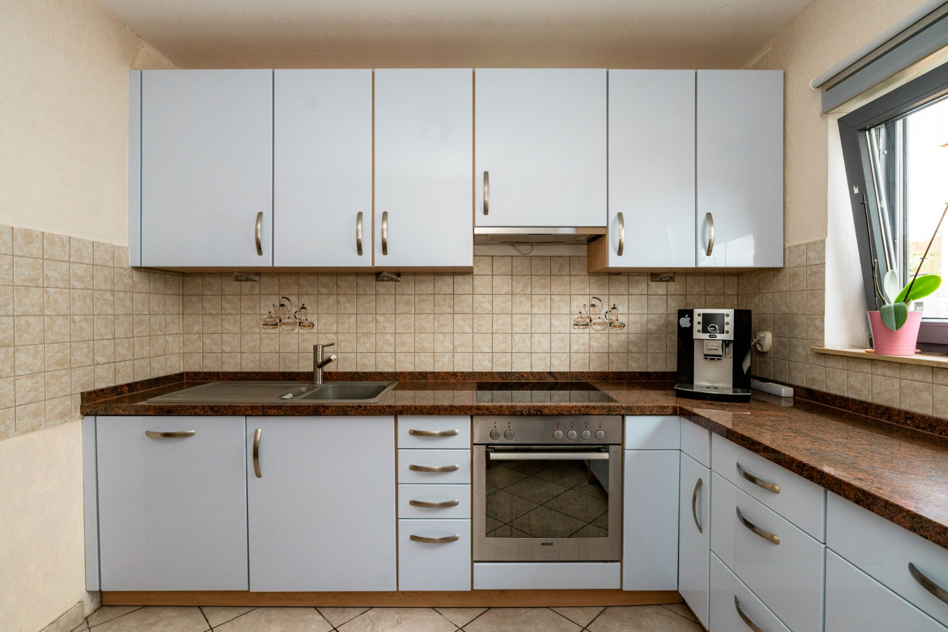 Moderne Einbauküche mit weißen Schränken, Elektroherd, Spüle und Kaffeemaschine am Fenster.