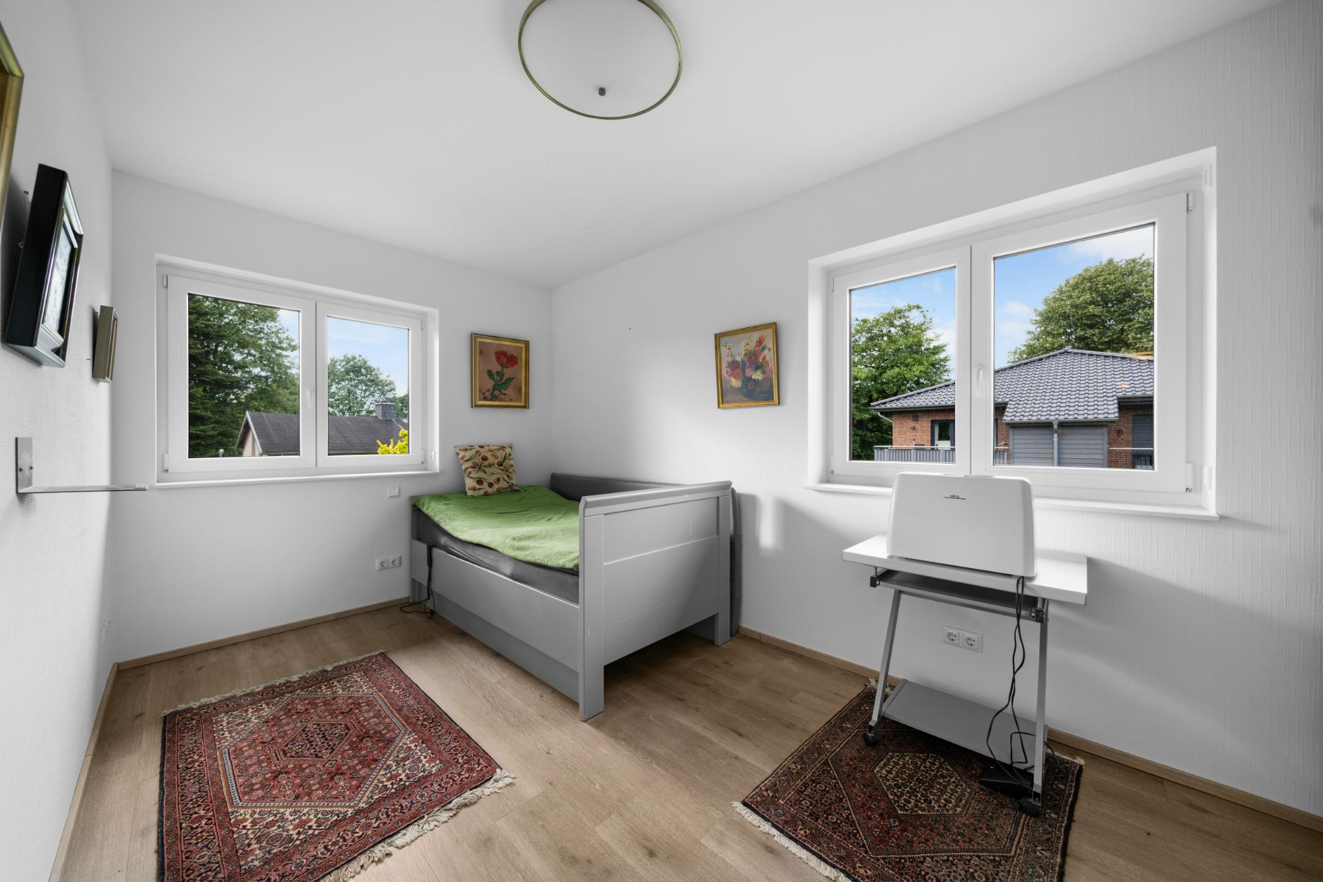 Helles Schlafzimmer mit Einzelbett, Schreibtisch, Teppichen und zwei Fenstern.