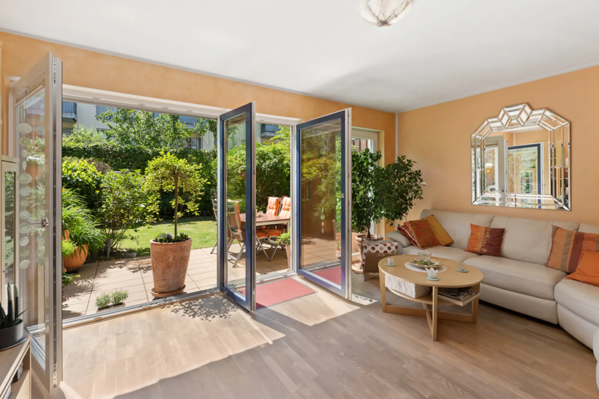 Helles Wohnzimmer mit offener Tür zur Terrasse und Garten, Sofa, Couchtisch und Spiegel an der Wand.