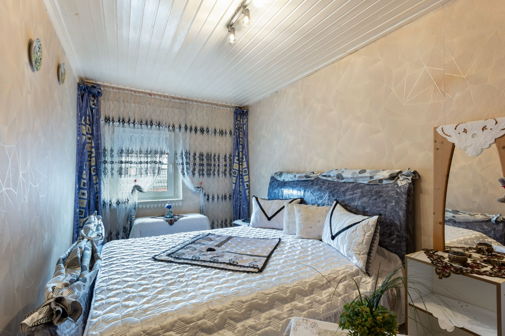 Kleines Schlafzimmer mit Doppelbett, Spiegel, Fenster mit Vorhängen und dekorierten Wänden.