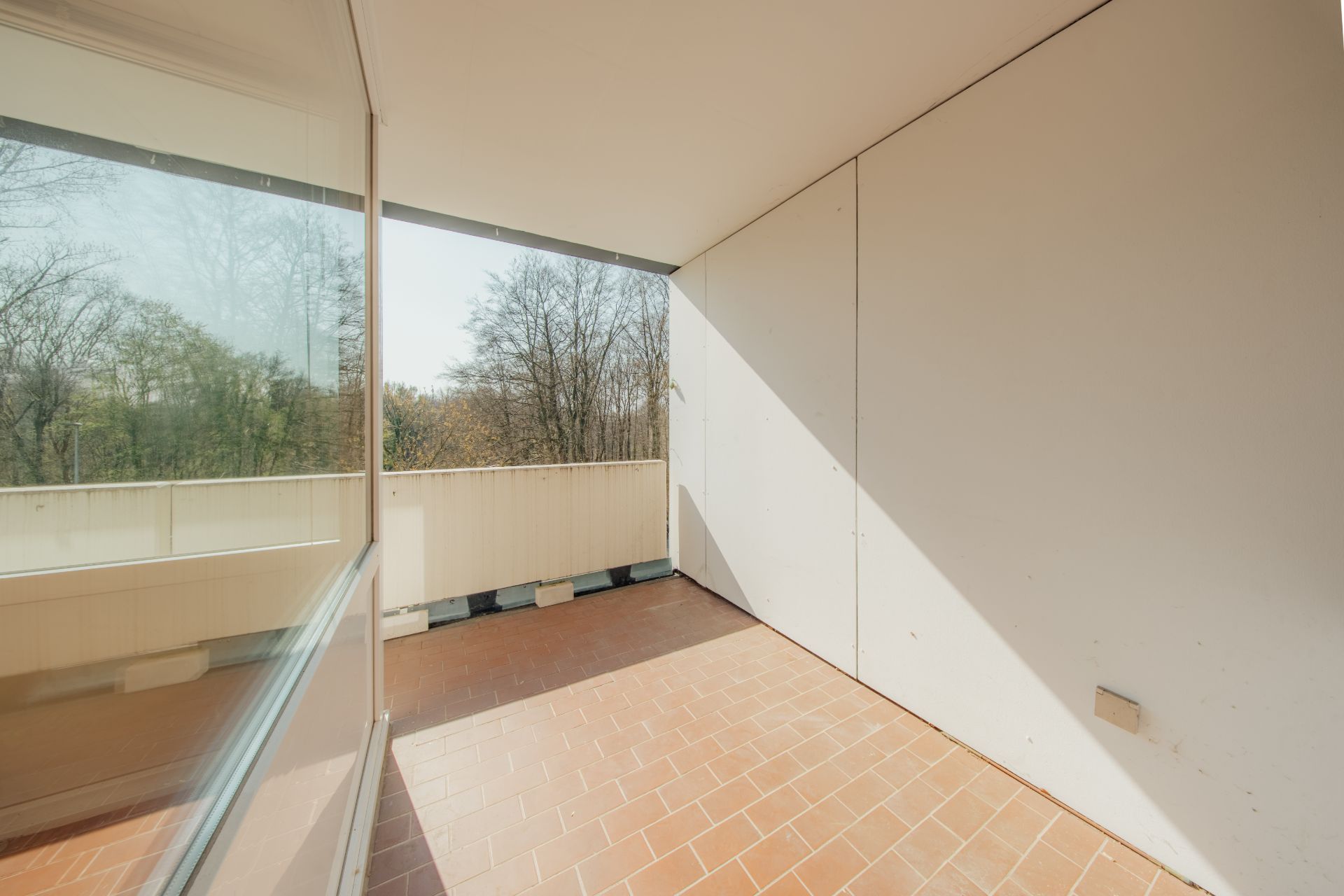 Überdachter Balkon mit braunem Fliesenboden und Blick auf Bäume.