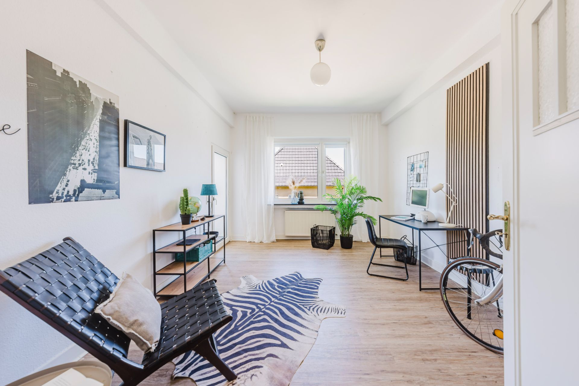 Modernes, helles Arbeitszimmer mit Schreibtisch, Stuhl, Sessel, Regal, Pflanzen und großem Fenster.