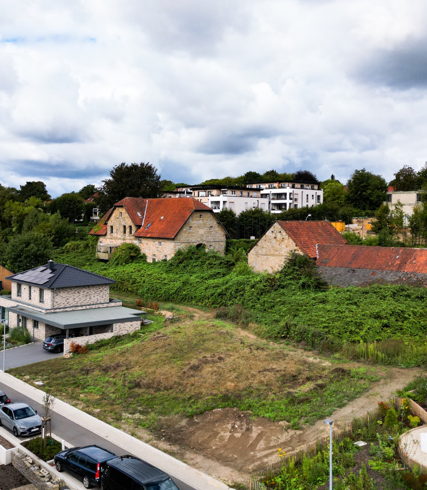 Wohnhäuser mit roten Dächern, grünes Gebüsch und parkende Autos an einer Straße.