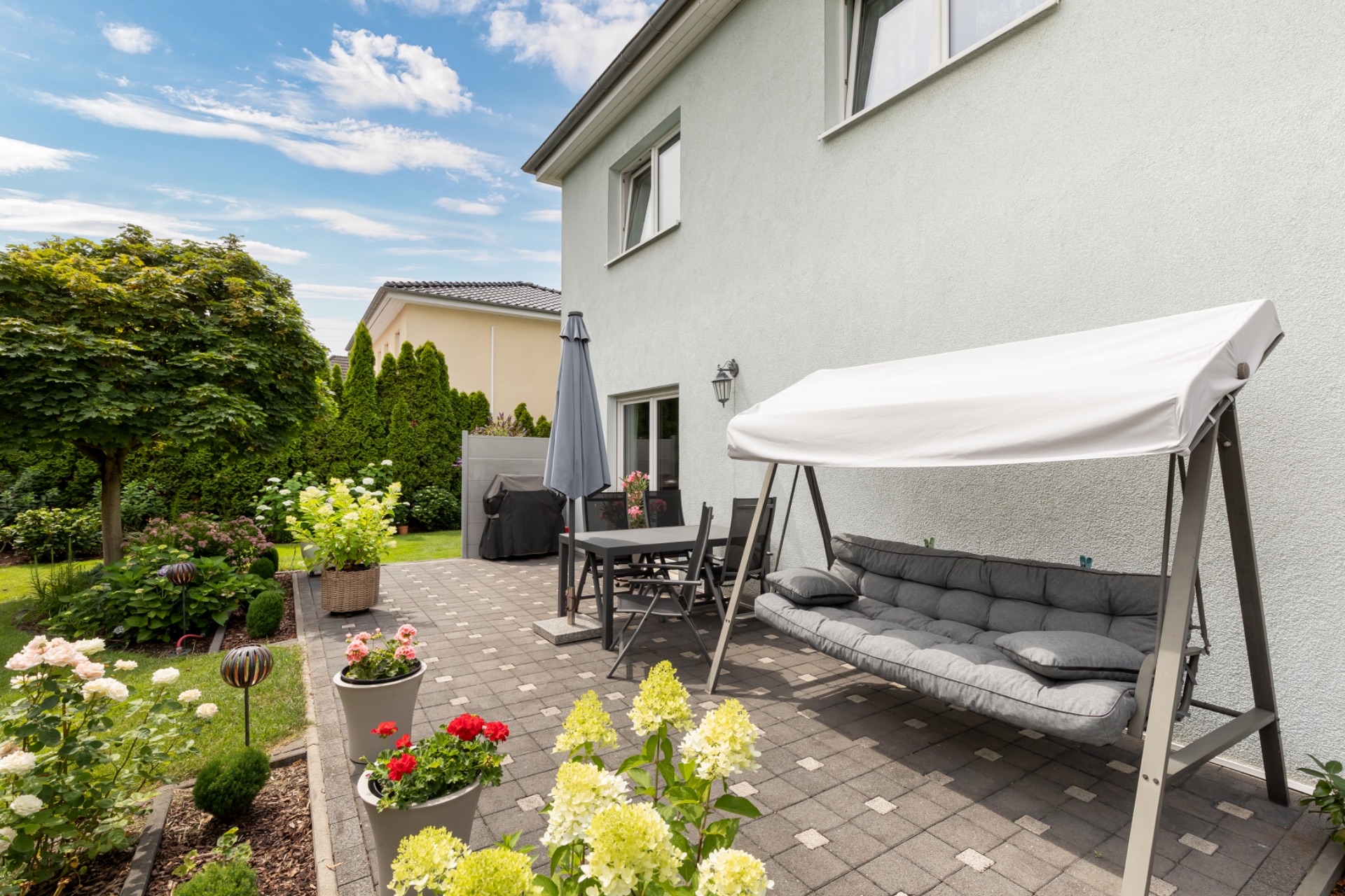 Gepflasterte Terrasse mit Hollywoodschaukel, Esstisch, Sonnenschirm und Blumen neben Wohnhaus.
