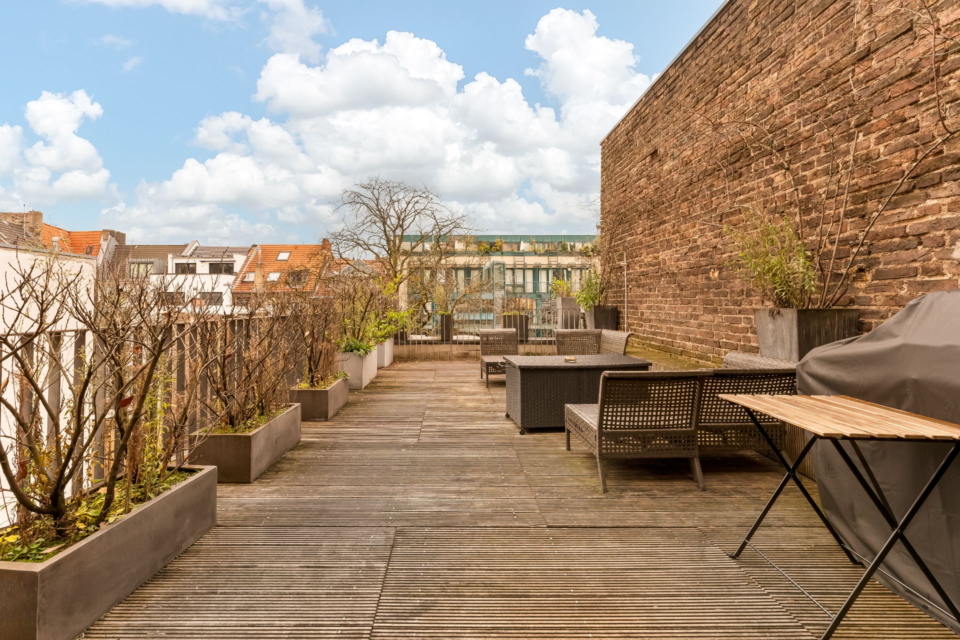 Holzterrasse mit Sitzgelegenheiten, Grill und Pflanzkübeln an einer Backsteinmauer.