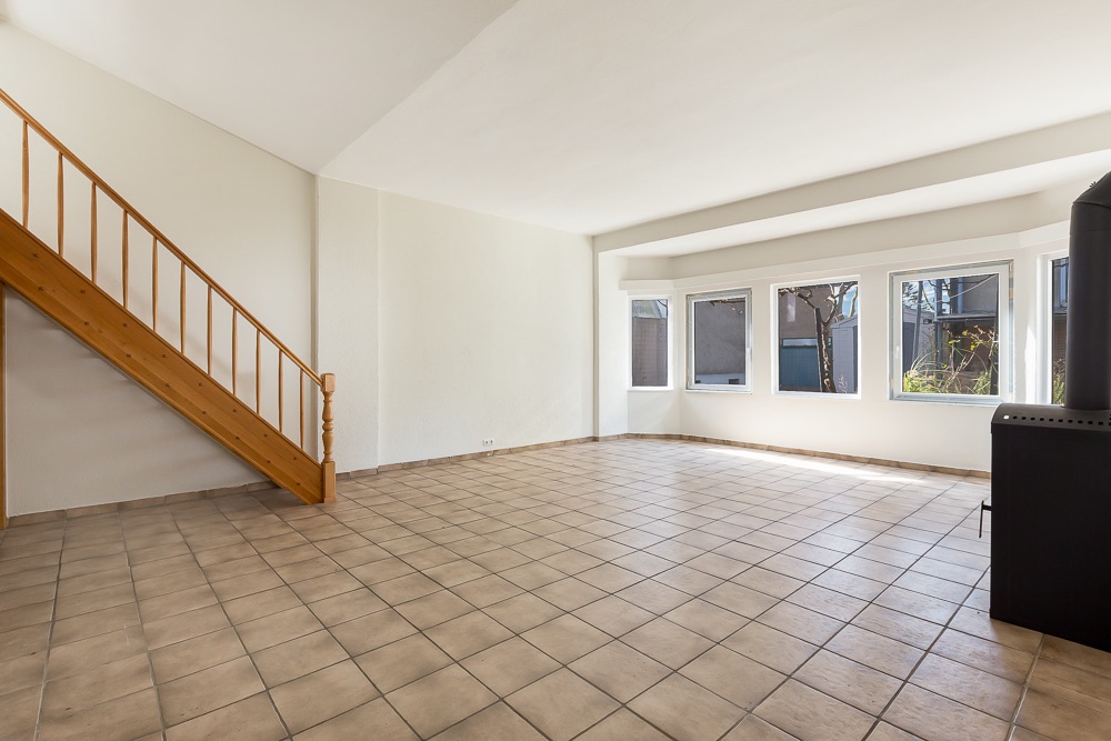 Heller Raum mit Fliesenboden, Holztreppe und großen Fenstern zur rechten Seite.
