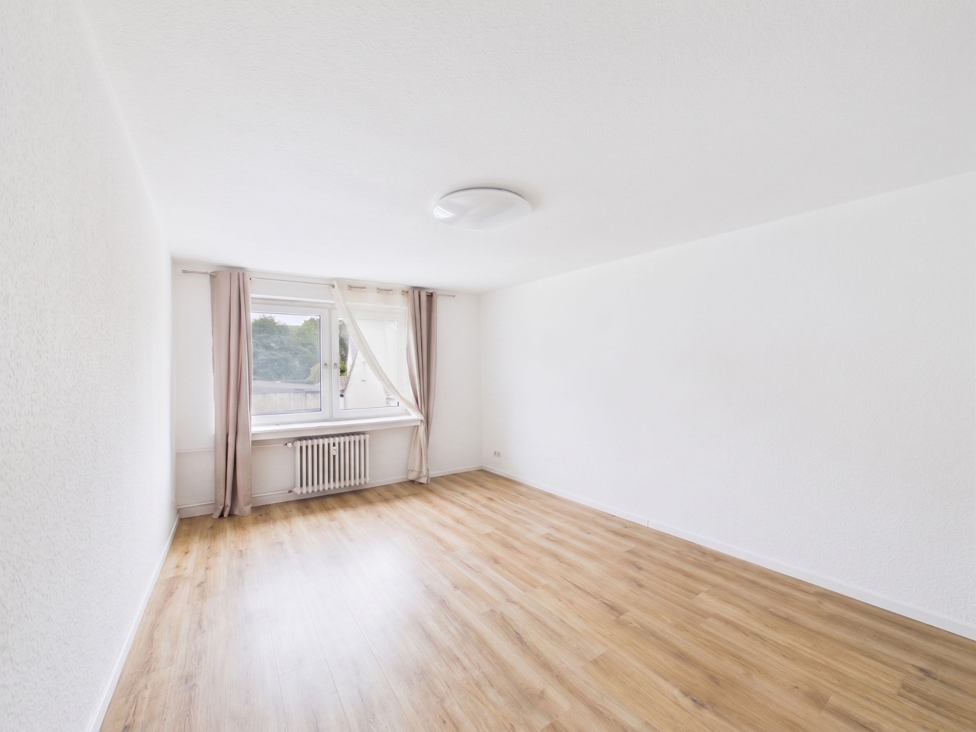Helles, leeres Zimmer mit Holzboden, weiß gestrichenen Wänden und einem Fenster mit Vorhängen.