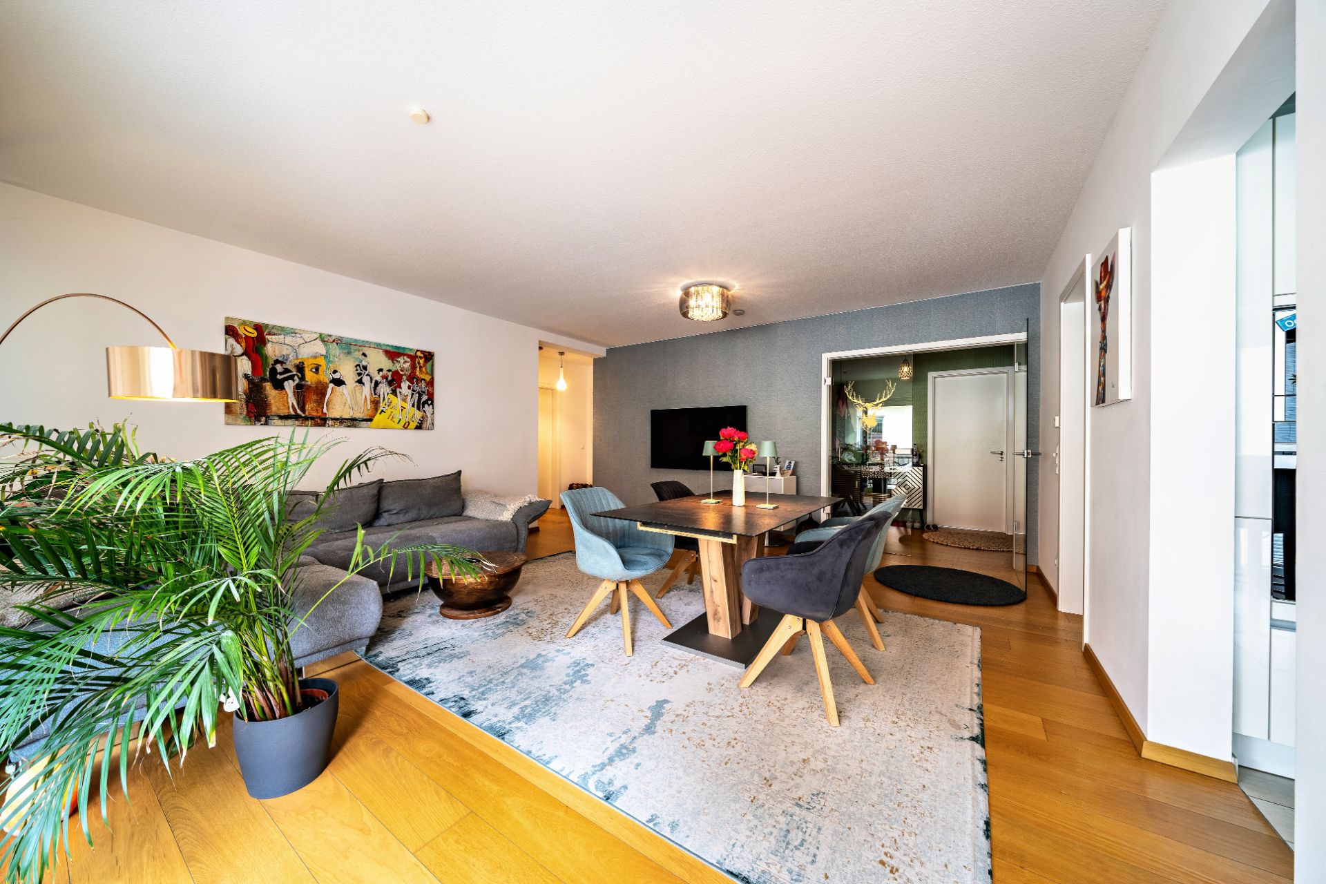 Modernes Wohnzimmer mit grauem Sofa, Esstisch, Stühlen, Teppich und grüner Zimmerpflanze.