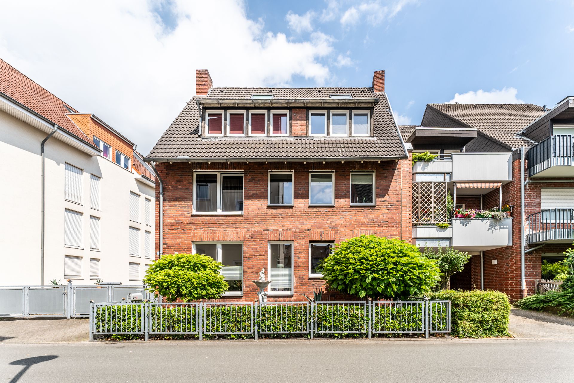 Mehrfamilienhaus aus rotem Backstein mit Vorgarten, Zaun und mehreren Fenstern.