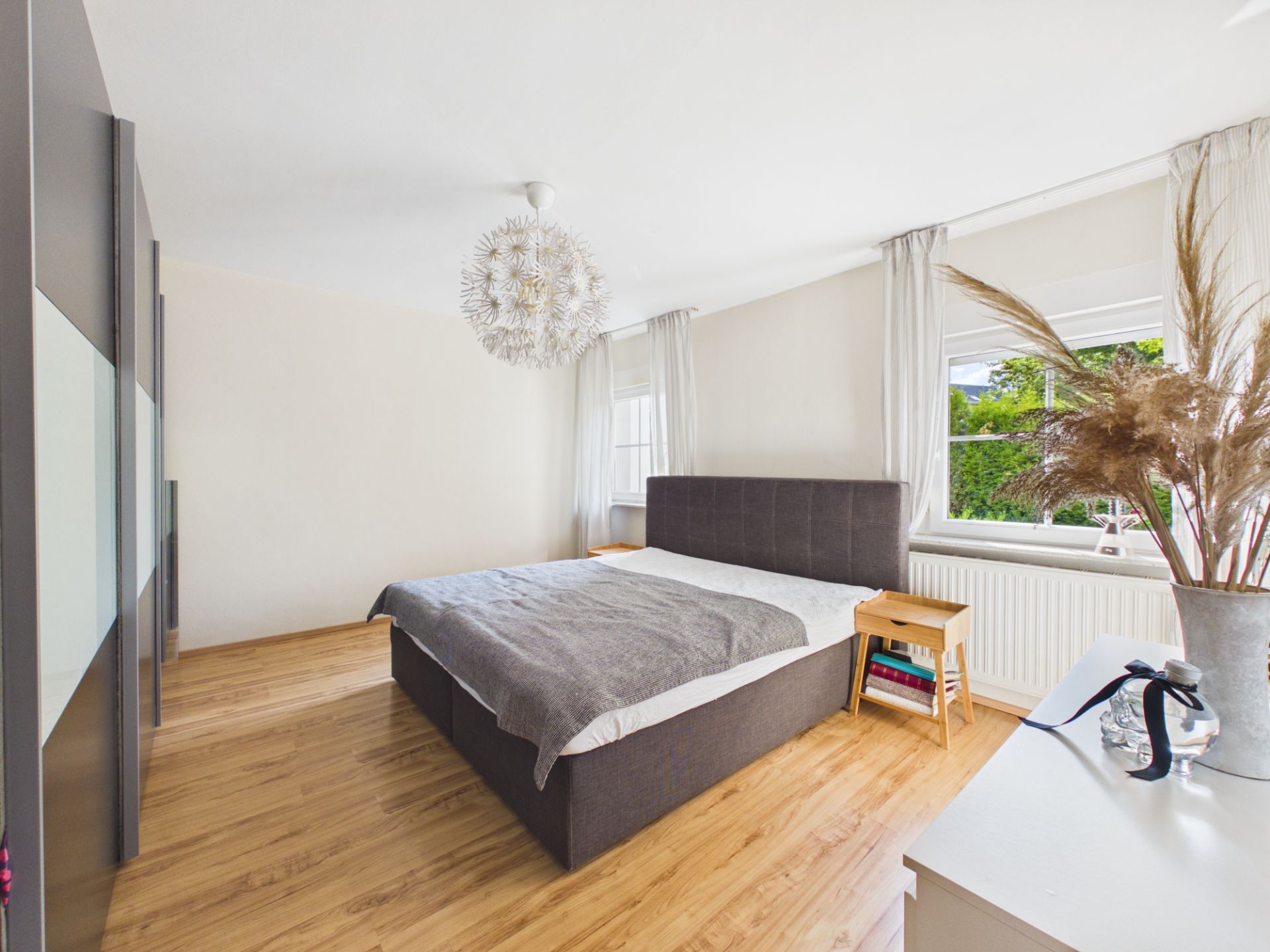 Helles Schlafzimmer mit großem Bett, Kleiderschrank, Nachttisch und zwei Fenstern.