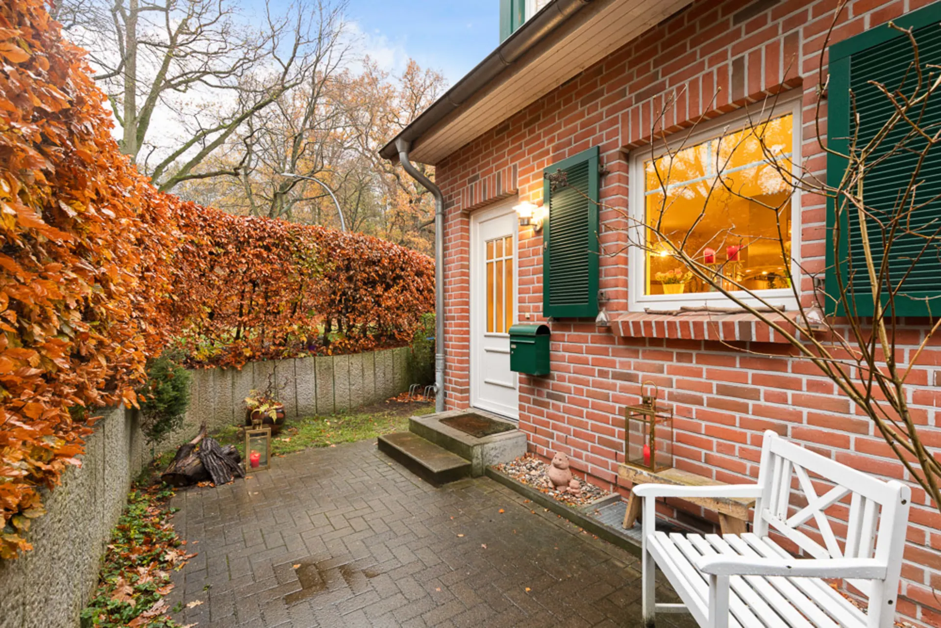 Gepflasterter Hauseingang mit weißer Bank, Backsteinfassade und grünen Fensterläden, umgeben von Hecke.