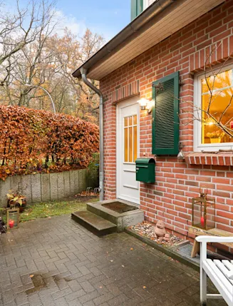 Gepflasterter Hauseingang mit weißer Bank, Backsteinfassade und grünen Fensterläden, umgeben von Hecke.