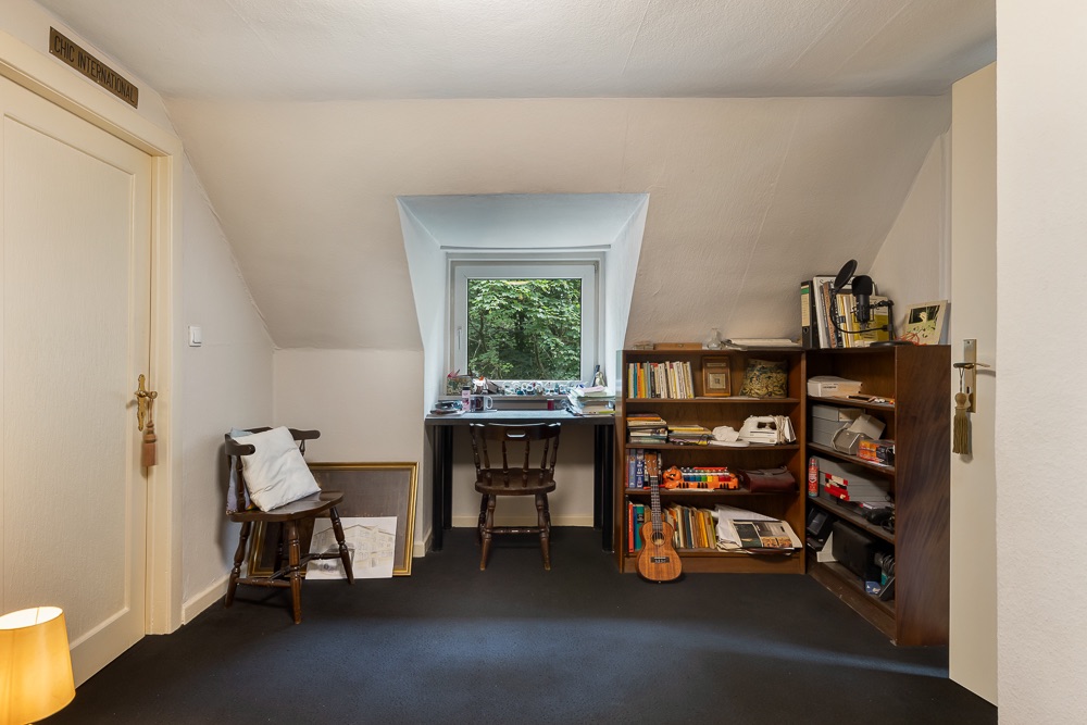 Kleines Arbeitszimmer mit Schreibtisch am Fenster, Stuhl und Bücherregalen mit Büchern und Gegenständen.