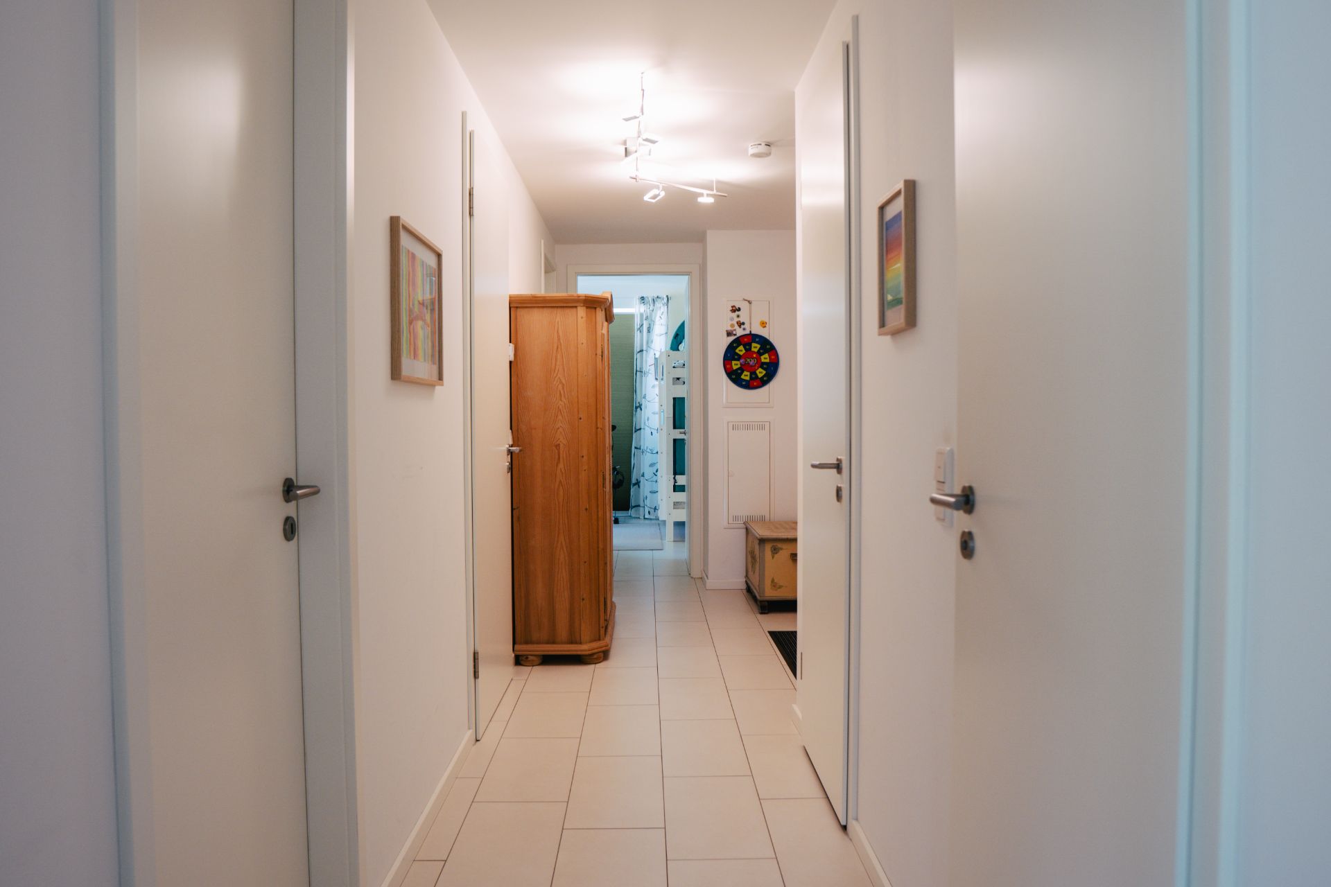 Heller Flur mit Fliesenboden, mehreren Türen, Bildern an den Wänden und einem Holzschrank am Ende.
