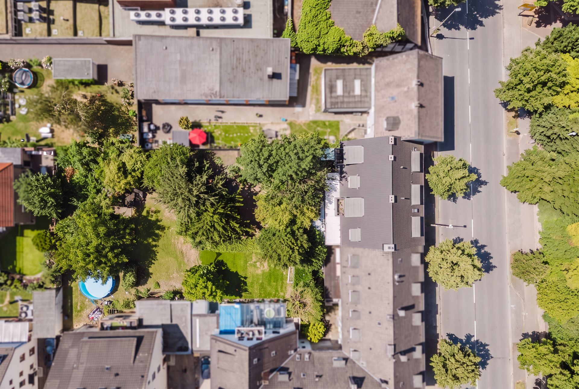 Luftaufnahme eines Wohngebiets mit Häusern, Bäumen, Straße und Gärten, darunter ein runder Pool.