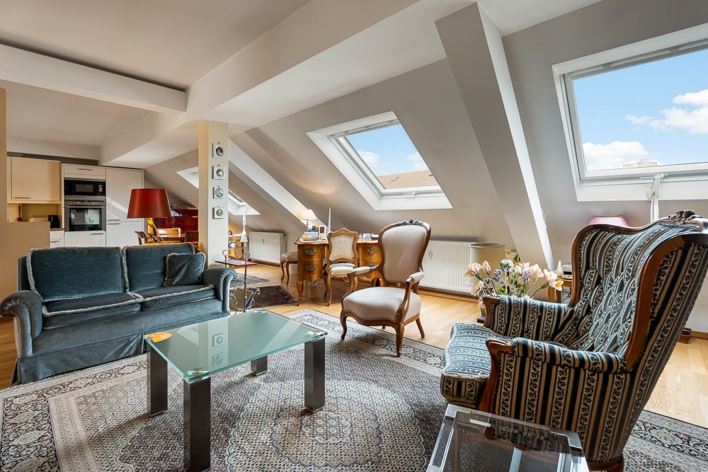 Wohnzimmer mit Dachfenstern, Sofa, Sesseln, Glastisch und Schreibtisch auf einem Teppich.