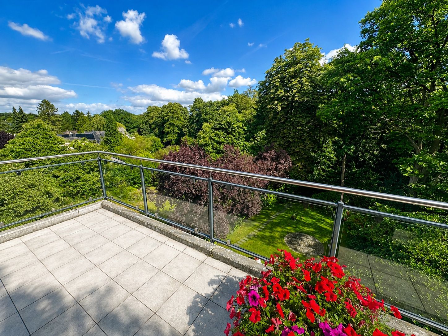 Geräumiger Balkon mit Glasgeländer und Blumen, Blick auf grünen Garten und Bäume unter blauem Himmel.
