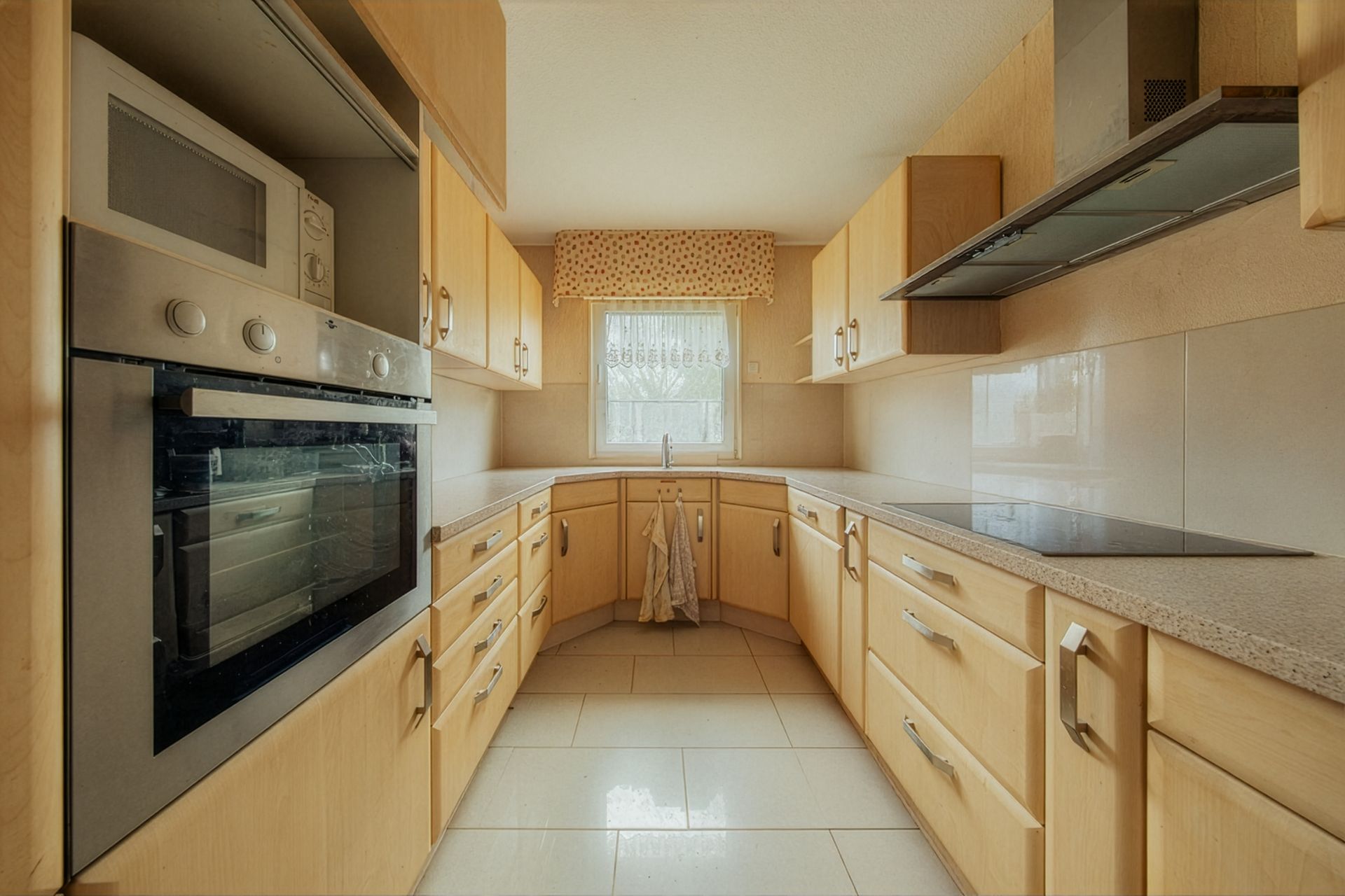 Helle Küche mit Holzschränken, Backofen, Herd, Fenster und gefliestem Boden.
