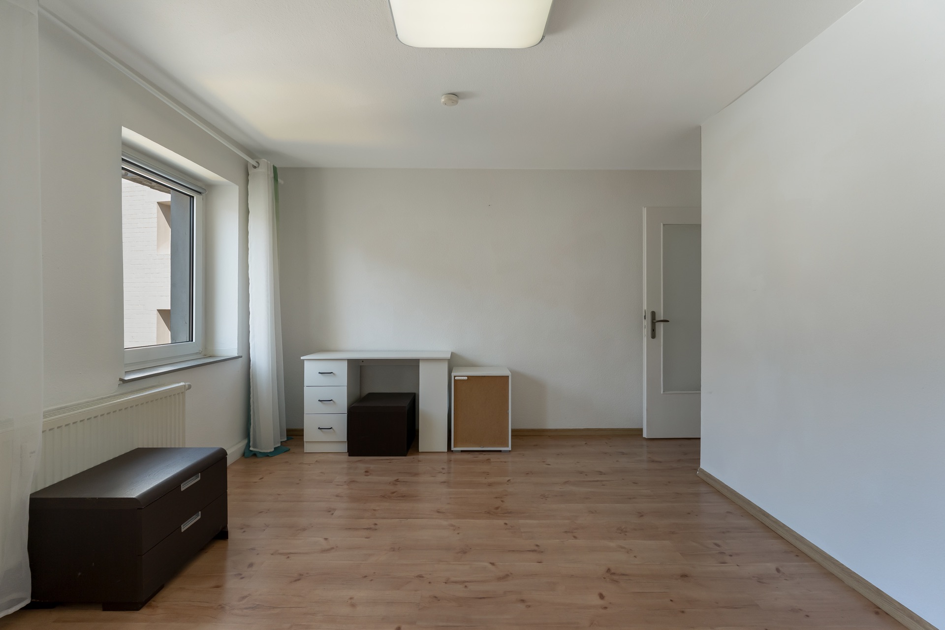 Helles Zimmer mit Holzboden, Fenster, kleinem Schreibtisch und Truhe.