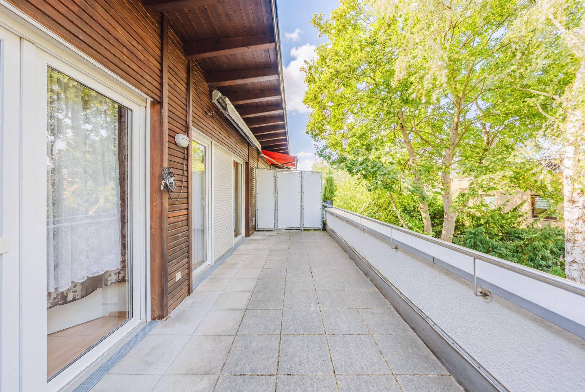 Großer Balkon mit Fliesenboden, Holzverkleidung und Glas-Schiebetüren, umgeben von Bäumen.