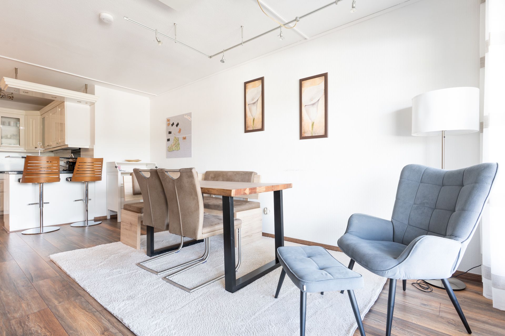 Helles Wohnzimmer mit Esstisch, Stühlen, Sessel, Hocker und Stehlampe auf Holzfußboden.