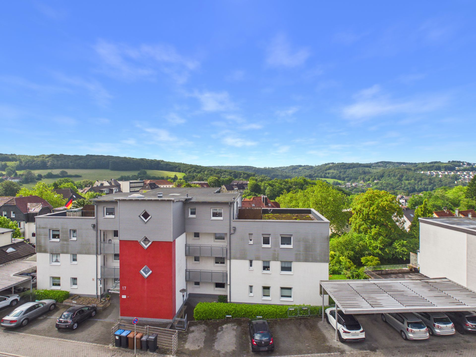 Mehrfamilienhaus mit rotem Mittelteil und Parkplätzen davor, im Hintergrund grüne Hügel.