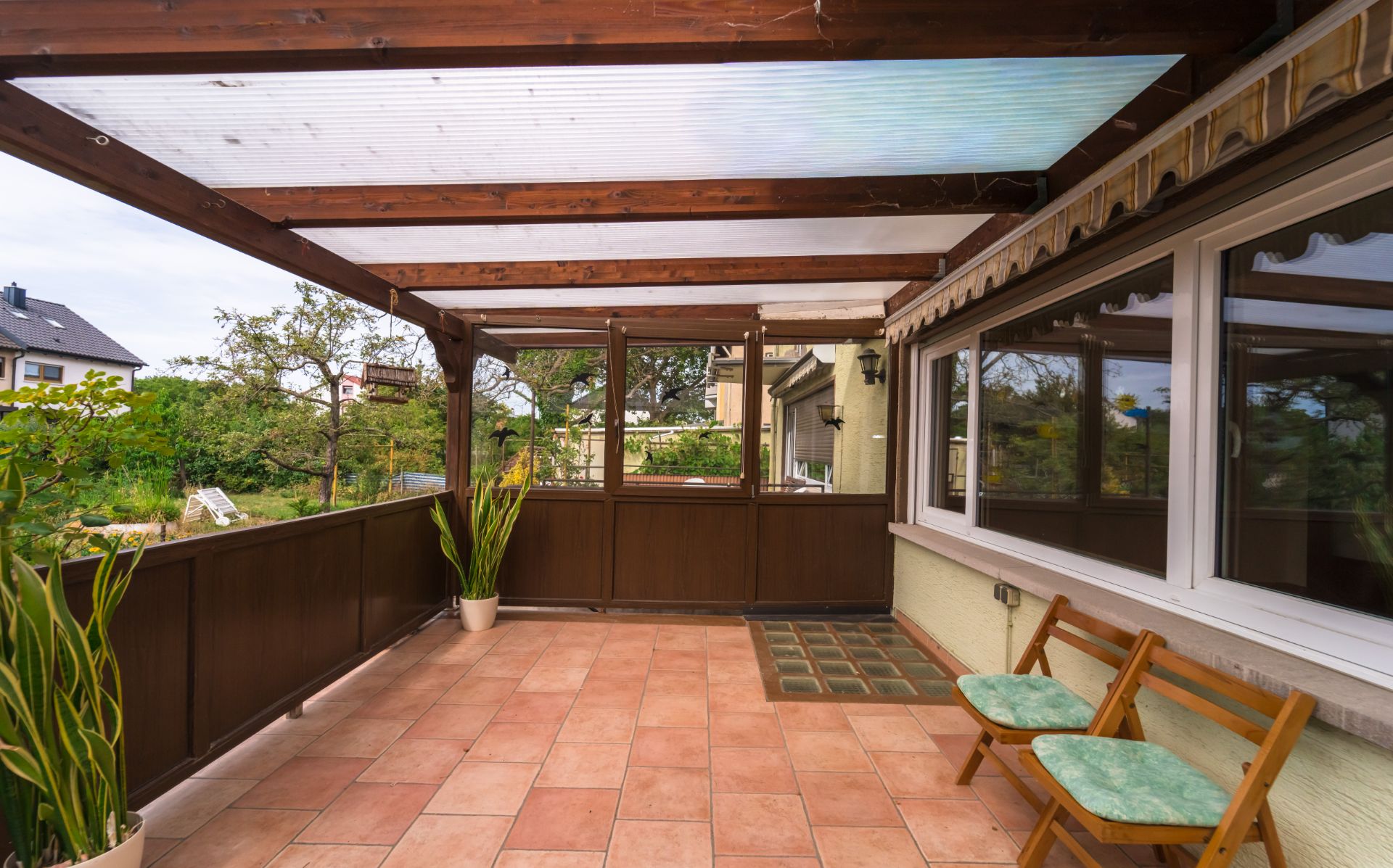 Überdachte Terrasse mit Fliesenboden, zwei Stühlen, Pflanzen und Blick in den Garten.