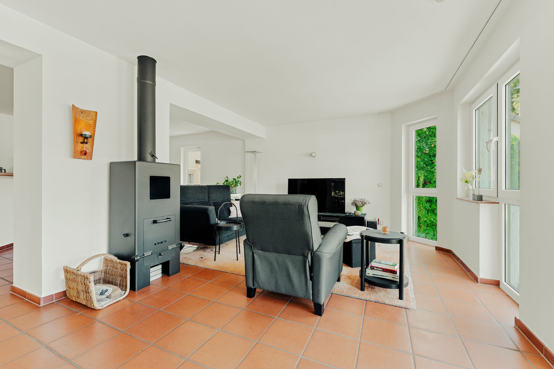 Helles Wohnzimmer mit Kaminofen, schwarzen Sesseln, Fernseher und großen Fenstern.