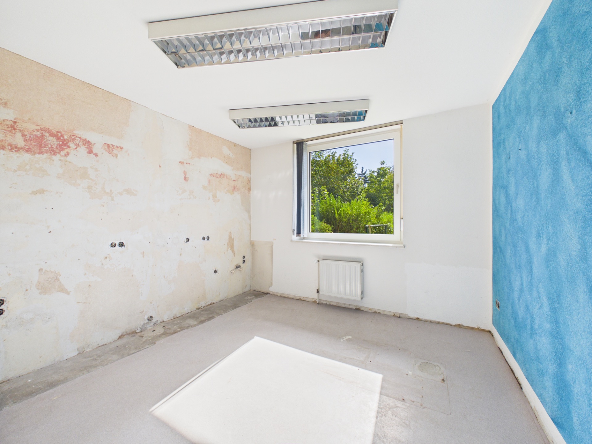 Leerer Raum mit abgenutzten Wänden, Fenster mit Blick auf Garten und zwei Deckenlampen.