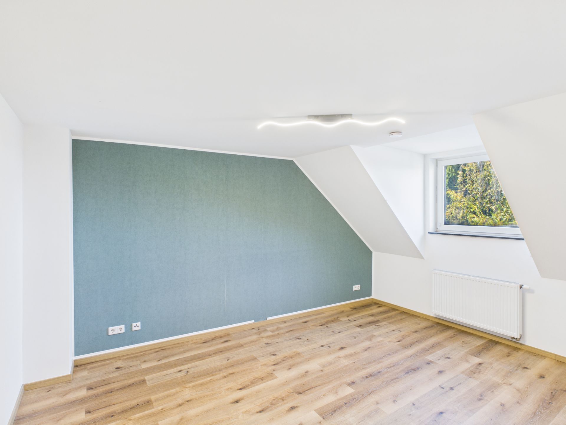 Helles, leeres Zimmer mit schräger Decke, Holzfußboden und Fenster zur rechten Seite.