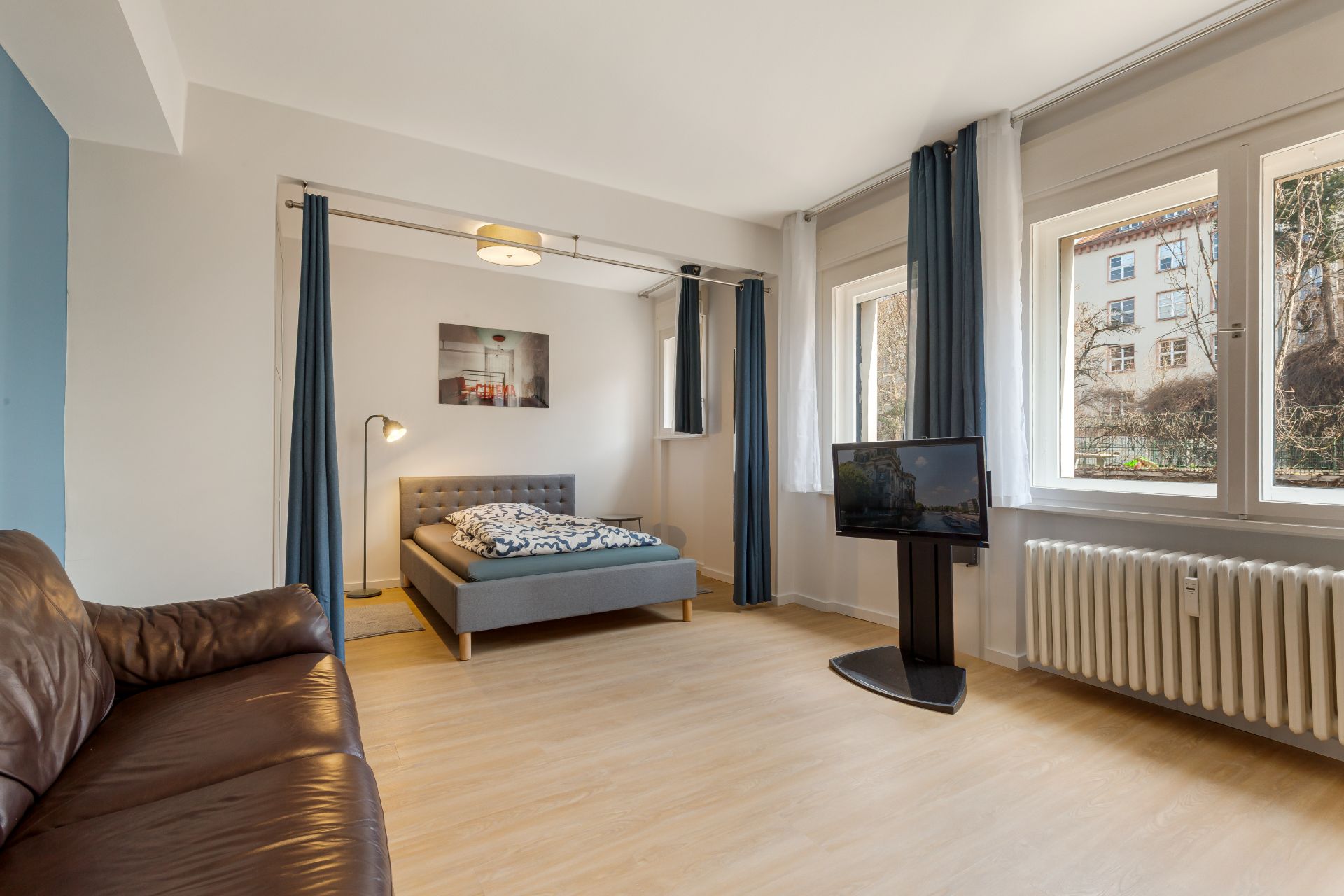 Helles Zimmer mit Bett, braunem Sofa, Fernseher und drei großen Fenstern mit Vorhängen.