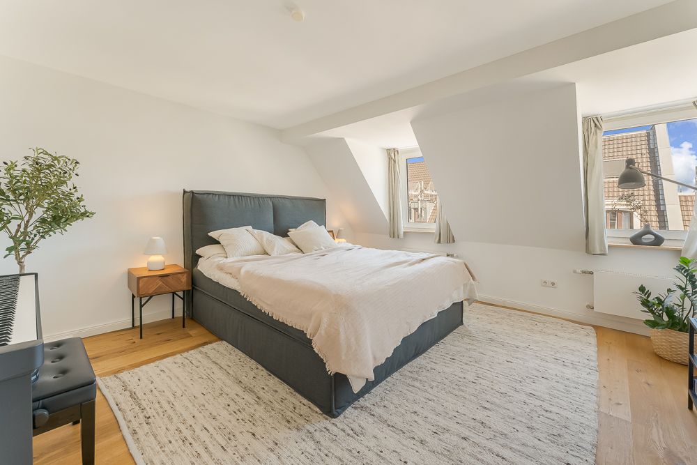 Helles Schlafzimmer mit Doppelbett, Teppich, Nachttischen, Pflanzen und zwei Fenstern.