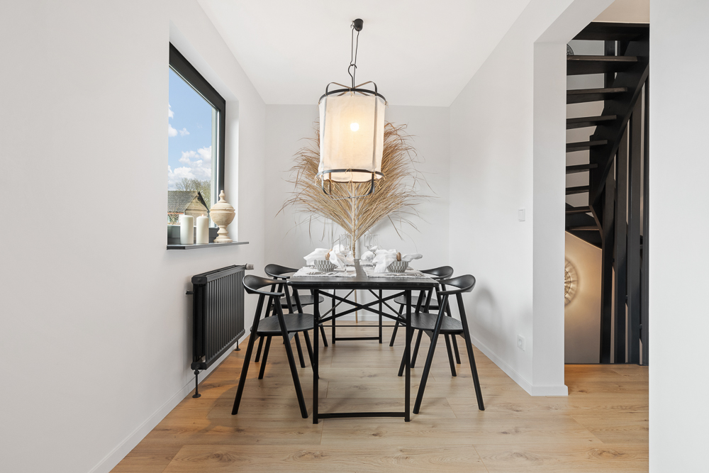 Heller Essbereich mit schwarzem Tisch, sechs Stühlen, großer Lampe und Treppe im Hintergrund.