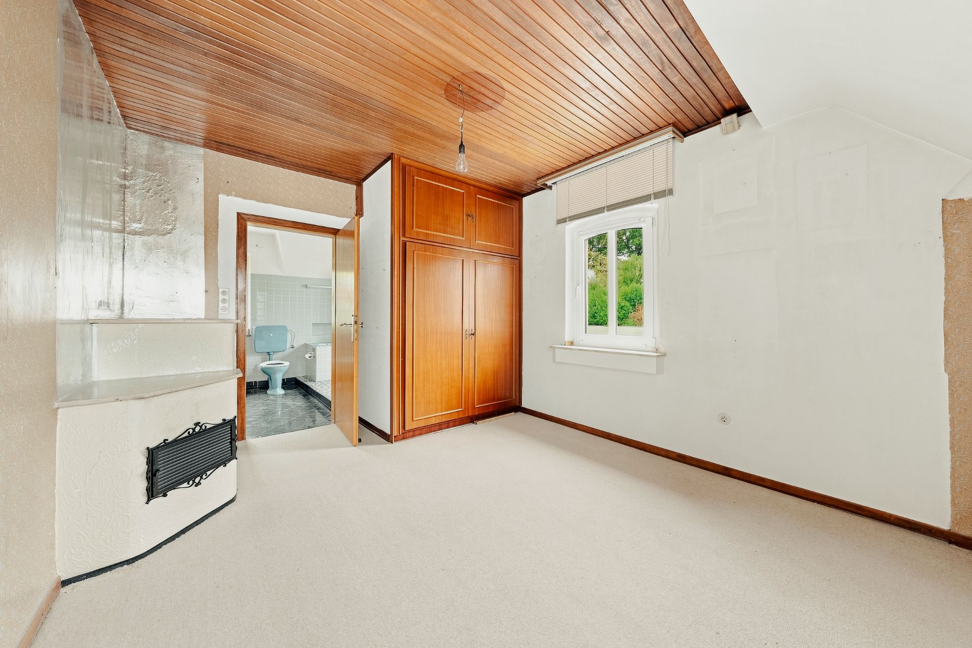 Leeres Zimmer mit Teppichboden, Holzdecke, Einbauschrank und Fenster. Offene Tür zeigt Badezimmer.