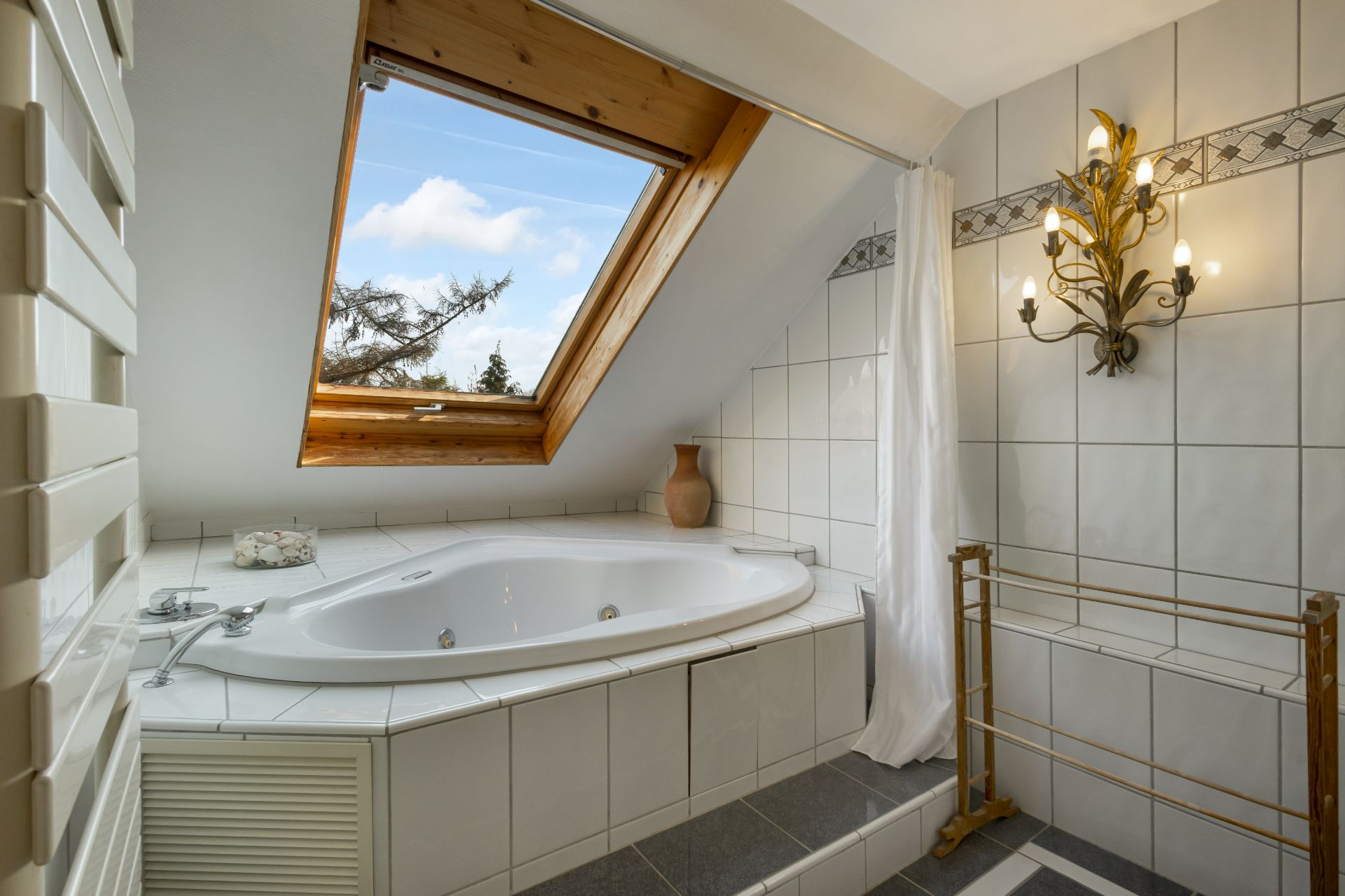 Badezimmer mit großer Eckbadewanne unter einem Dachfenster und dekorativer Wandlampe.