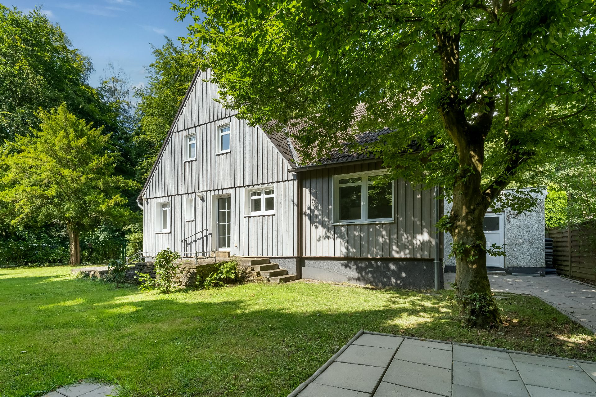 Grau gestrichenes Einfamilienhaus mit Garten, Bäumen und gepflastertem Weg bei sonnigem Wetter.