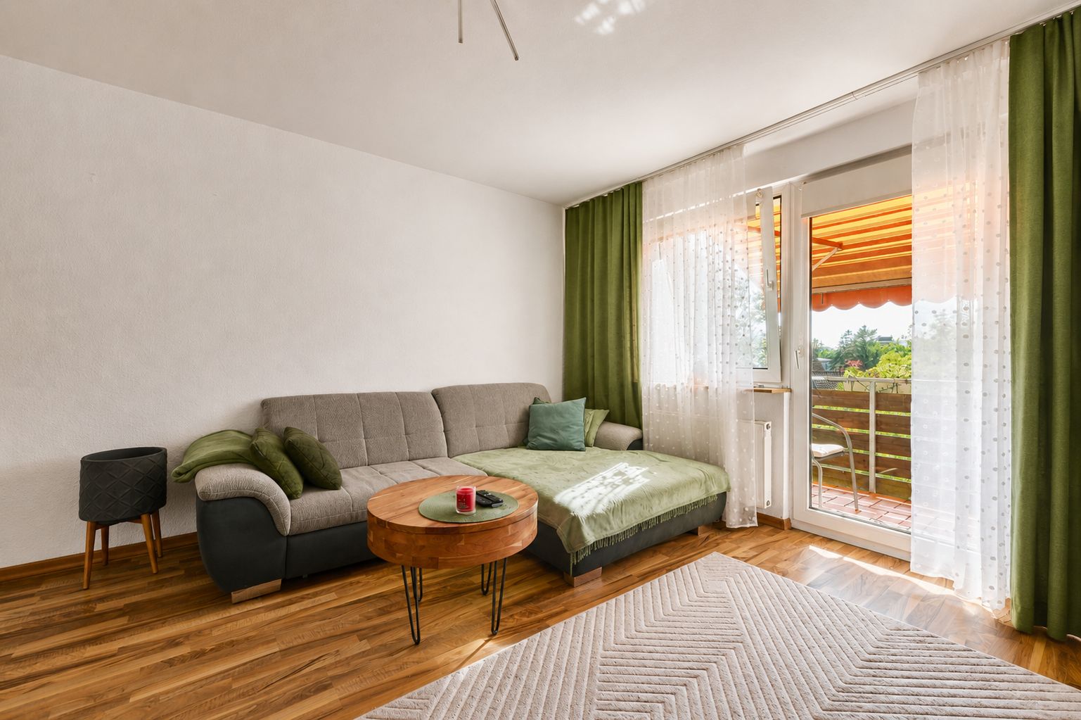 Wohnzimmer mit grauem Sofa, grünem Teppich, rundem Tisch und Zugang zu einem Balkon mit Vorhängen.