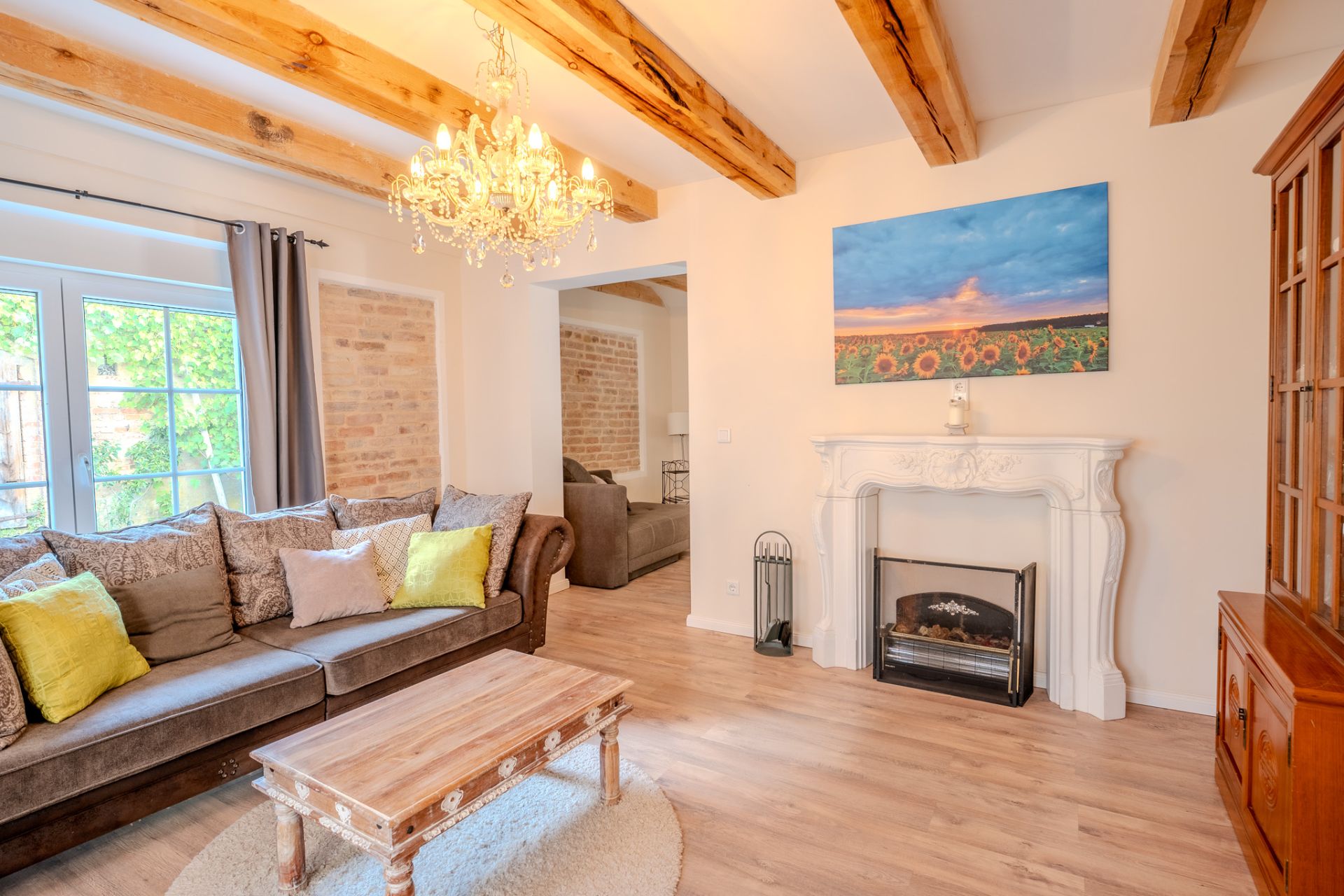 Helles Wohnzimmer mit Sofa, Holztisch, Kamin, Kronleuchter und offenem Zugang zu weiterem Raum.