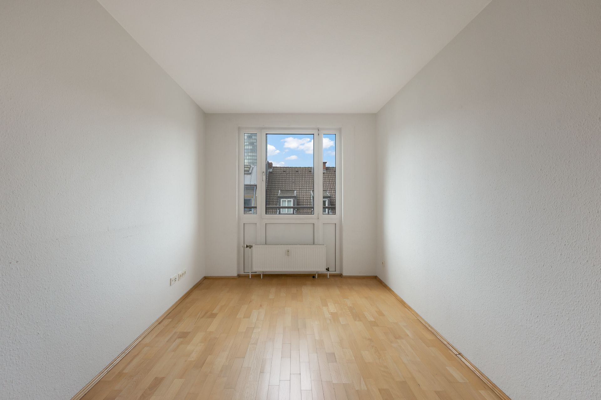 Leerer Raum mit hellem Holzboden, weißen Wänden und Fenster mit Heizung darunter.