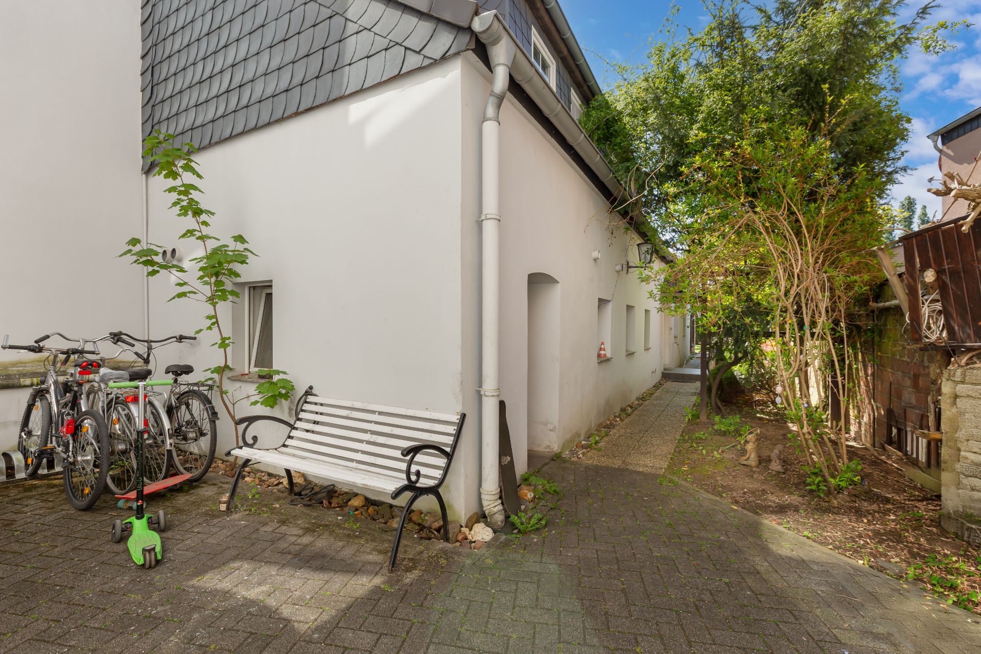 Innenhof mit Bank, mehreren Fahrrädern, einem Kindertretroller und Bäumen neben einem weißen Gebäude.