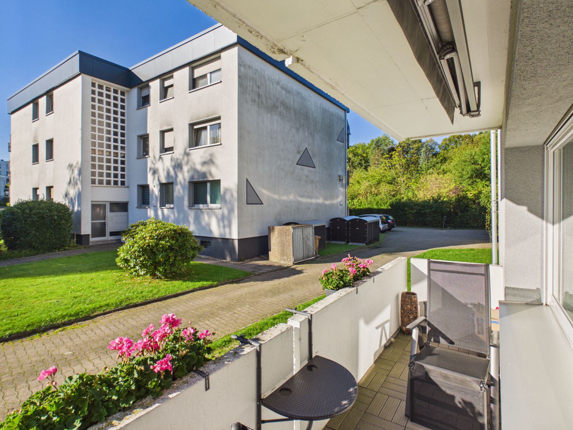 Blick von einem Balkon mit Stuhl und Tisch auf einen Hof mit Wohnhaus und Grünfläche.