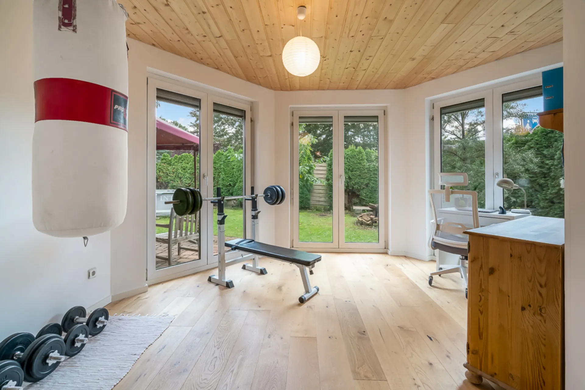 Heller Raum mit Boxsack, Hanteln, Trainingsbank und Schreibtisch mit Stuhl vor großen Fenstern.