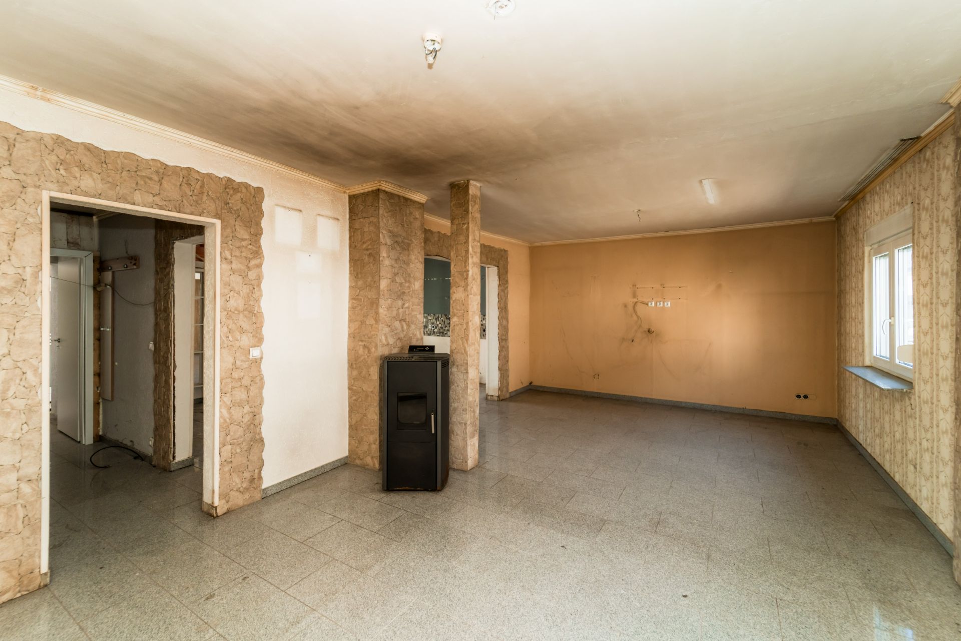 Leerer Wohnraum mit großen Fenstern, sichtbaren Türen und einem Ofen in der Mitte.
