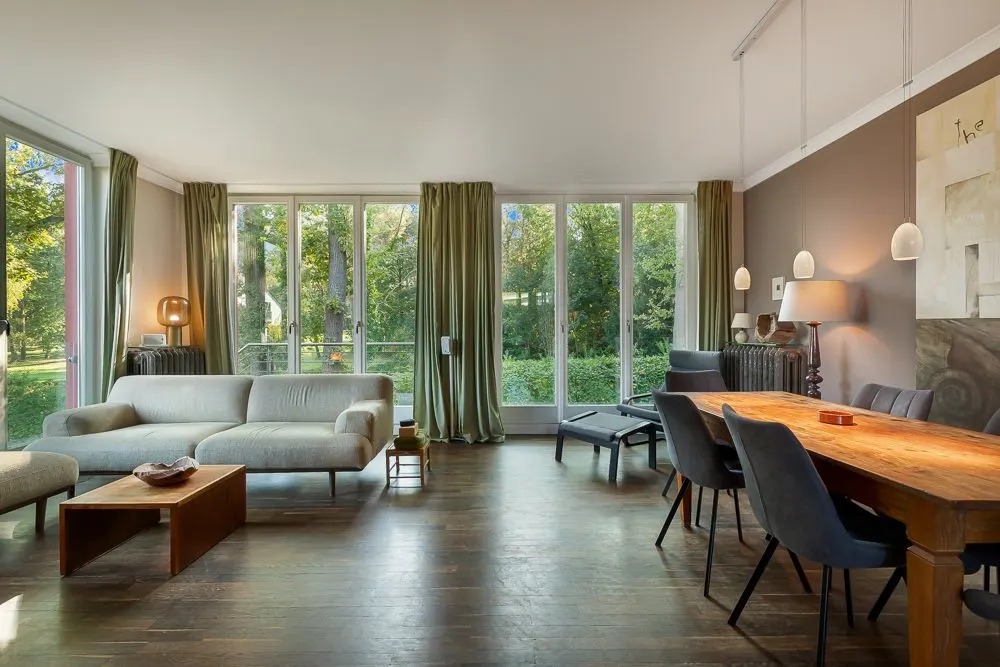 Helles Wohnzimmer mit großen Fenstern, Sofa, Esstisch und Stühlen auf Holzfußboden.