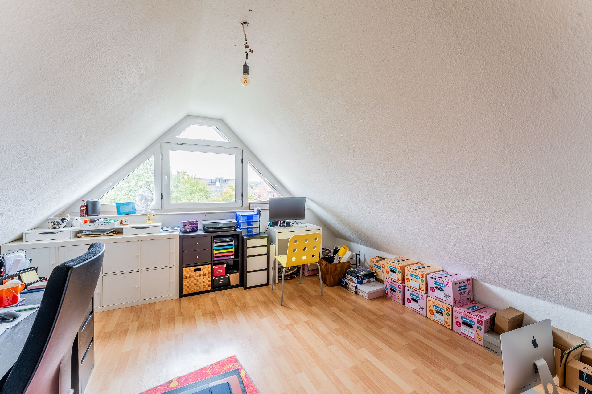 Dachzimmer mit Schreibtisch, Computer, Regalen und mehreren Kartons auf dem Boden.